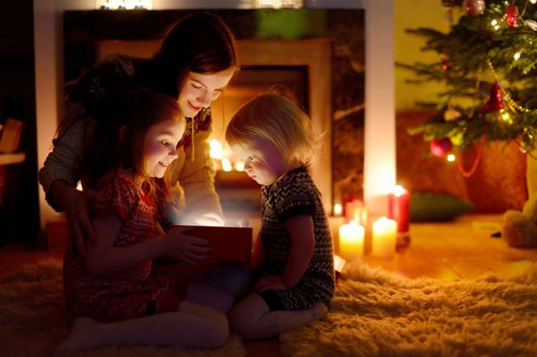 مادر جوان و دو دختر کوچکش در زمستان یک هدیه کریسمس جادویی توسط یک درخت کریسمس در اتاق نشیمن دنج باز می کنند
