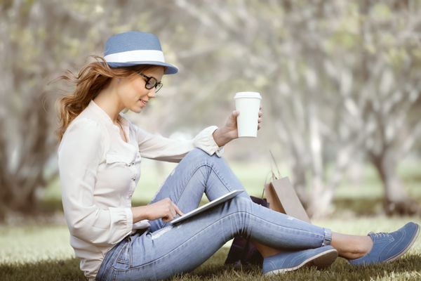 زن جوان شاد یک فنجان قهوه یکبار مصرف که روی چمن نشسته و از پد لمسی روی تبلت خود در پارک تابستانی استفاده می کند