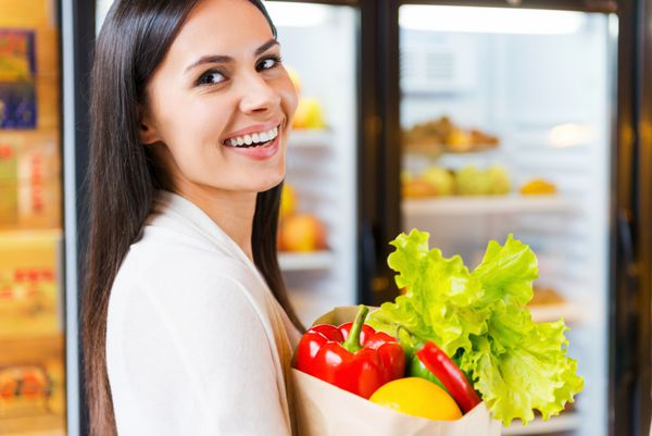 من محصولات ارگانیک را انتخاب می کنم زن جوان زیبا کیسه خرید با میوه ها در دست دارد و در حالی که در فروشگاه مواد غذایی نزدیک یخچال ایستاده لبخند می زند