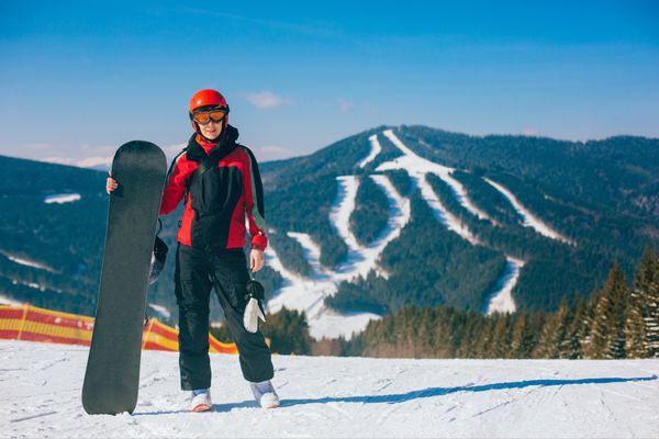 ورزش های زمستانی زن جوان اسنوبورد سوار در حال ژست گرفتن مقابل کوه با دامنه‌های پیست اسکی