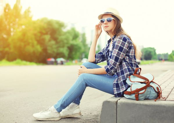 زن زیبا با عینک آفتابی کلاه حصیری و کوله پشتی که در تابستان در شهر بیرون از خانه نشسته است
