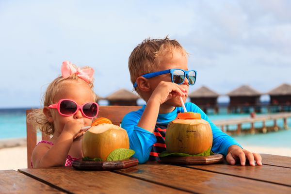 پسر کوچولو و دختر نوپا در حال نوشیدن دم نارگیل در استراحتگاه ساحلی گرمسیری سفرهای مجلل کودکان