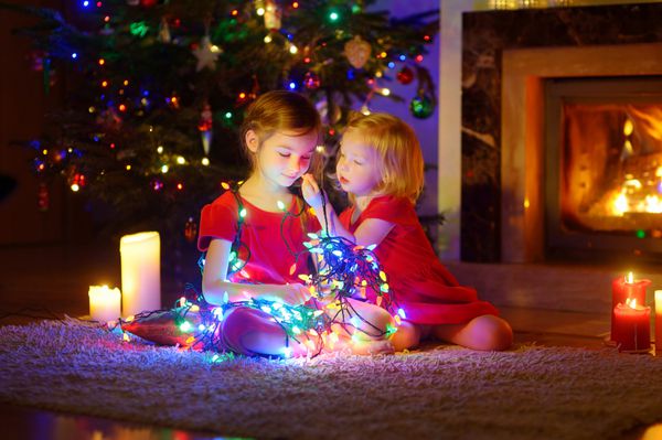 دختر بچه های شایان ستایش با چراغ های کریسمس در کنار یک شومینه در اتاق ناهار خوری تاریک و دنج