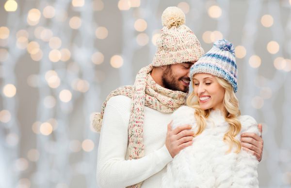 مفهوم زمستان مد زوج کریسمس و افراد - مرد و زن خندان با کلاه و روسری که روی پس زمینه چراغ های تعطیلات در آغوش گرفته اند