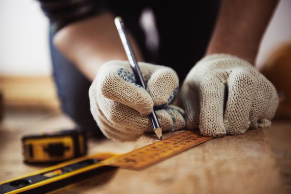 نمای نزدیک از دست های صنعتگر در دستکش های محافظ اندازه گیری تخته چوبی با خط کش و مداد مفهوم کارهای چوبی و بازسازی