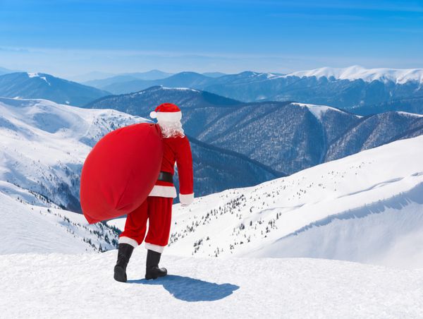 بابا نوئل با کیف کریسمس پر از هدایا در برابر منظره پیست اسکی کوهستانی زمستانی برفی و آسمان آبی کارت تبریک سال نو یا کریسمس