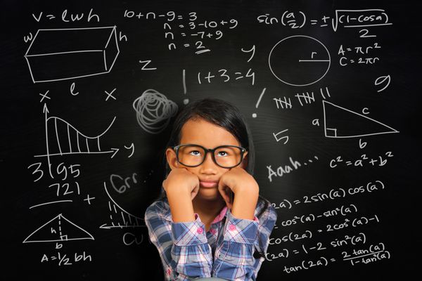 دختر دانشجوی آسیایی کوچک با عینکی که بی حوصله و خسته را روی تخته سیاه سبز رنگ که معادل های ریاضی روی آن نوشته شده نشان می دهد
