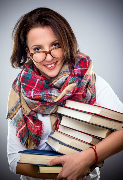 زن خوش استایل با روسری و عینک رنگارنگ زمستانی که انبوهی از کتاب‌های جلد گالینگور را زیر بغلش نگه داشته و به دوربین لبخند می‌زند
