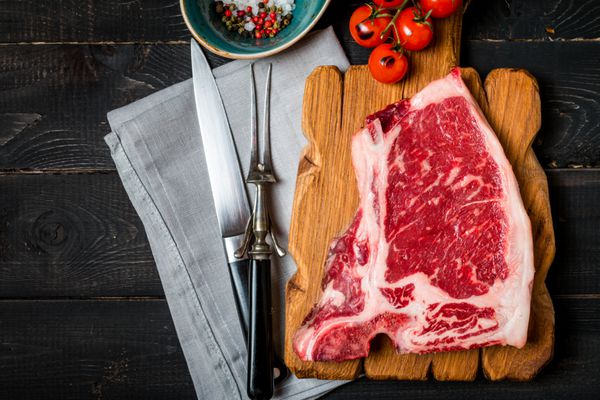 استیک کلوپ گوشت تازه خام چاشنی و چنگال گوشت و چاقو روی زمینه چوبی تیره نمای بالا
