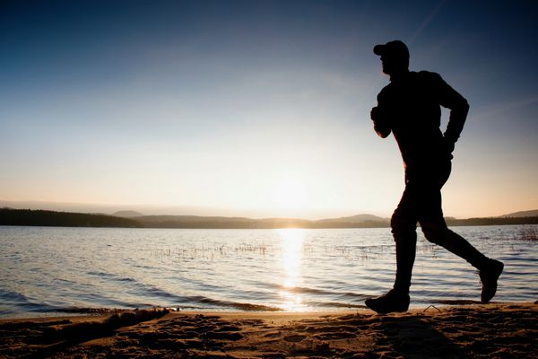 مرد دونده در ساحل ورزشکاری که با کلاه بیسبال می دود و در طلوع آفتاب بالای ساحل شنی می دود