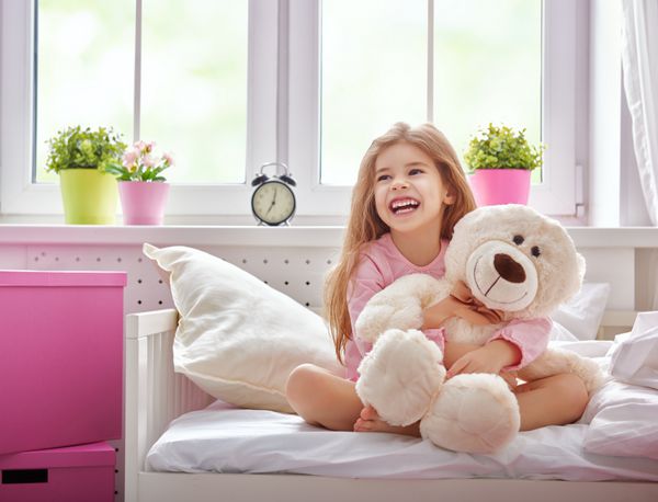 دختر بچه از خواب بیدار شد و از آفتاب صبح لذت می برد دختر می خندد و خرس عروسکی را در آغوش می گیرد