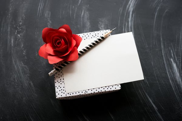 گل رز قرمز کاغذی و جعبه هدیه با یک کارت خالی روی تخته سیاه