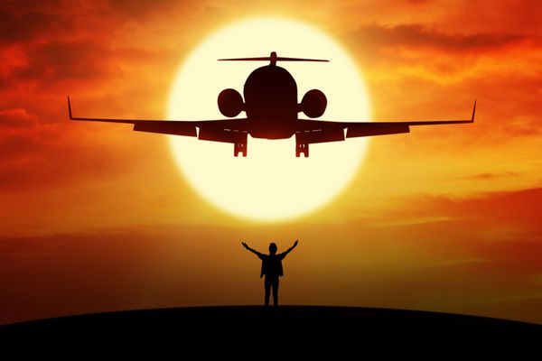 تصویر مرد جوانی که از آزادی لذت می برد در حالی که در هنگام غروب آفتاب روی تپه زیر یک هواپیمای در حال پرواز ایستاده است