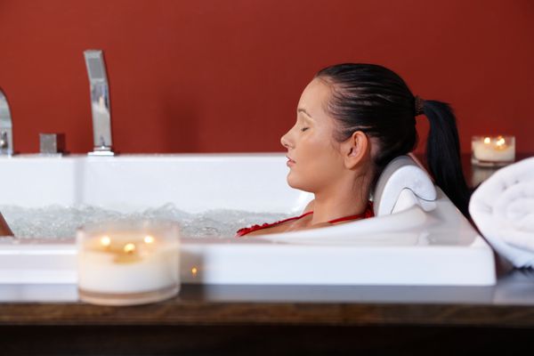 زن جوان در حال لذت بردن از حمام حباب سلامتی با چشمان بسته در نور شمع