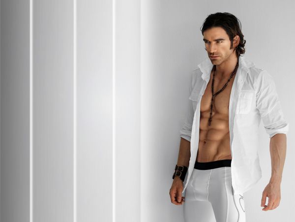 پرتره یک مدل تناسب اندام خوش تیپ با پیراهن سفید باز و شلوار بلند باکسر سفید در پس زمینه مدرن