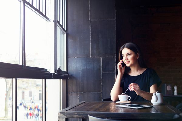 دختر جوان جذاب در حال صحبت با تلفن همراه و لبخند زدن در حالی که در اوقات فراغت در کافی شاپ تنها نشسته است زن خوشحال در حال استراحت در کافه سبک زندگی قهوه