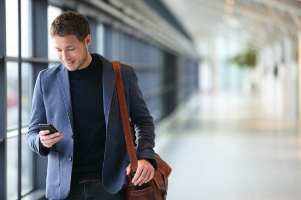 مرد در تلفن هوشمند - مرد جوان تجاری در فرودگاه تاجر حرفه‌ای شهری با استفاده از گوشی‌های هوشمند در حال لبخند زدن خوشحال در داخل ساختمان اداری یا فرودگاه مرد خوش تیپ با کت کت و شلوار در داخل خانه