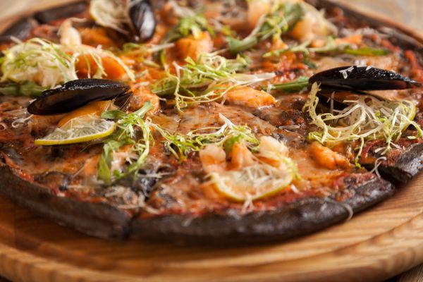 پیتزا ایتالیایی دی ماره با خمیر سیاه و غذاهای دریایی روی میز چوبی