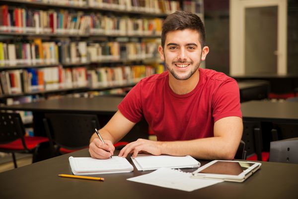 دانشجوی پسر جذاب دانشگاه در حال انجام تکالیف در کتابخانه مدرسه و لبخند زدن