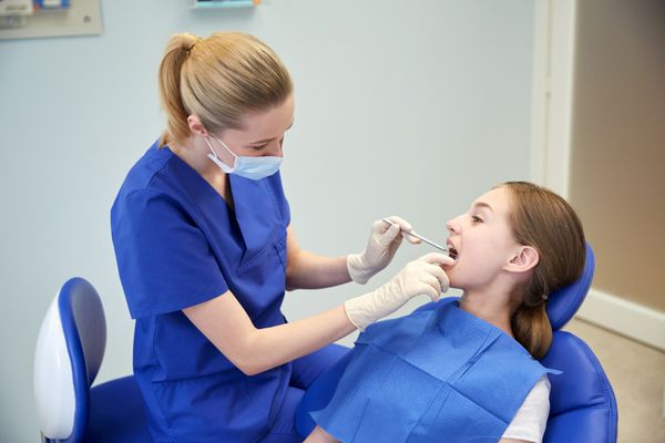 مفهوم مردم پزشکی دهان شناسی و مراقبت های بهداشتی - دندانپزشک زن شاد با آینه در حال بررسی دندان های دختر بیمار در مطب کلینیک دندانپزشکی