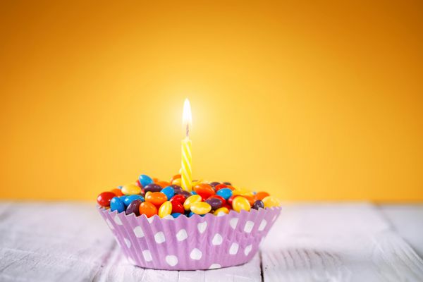 تزیین کاپ کیک تولد با یک شمع روشن و آب نبات های رنگارنگ روی زمینه زرد کارت تولد مبارک