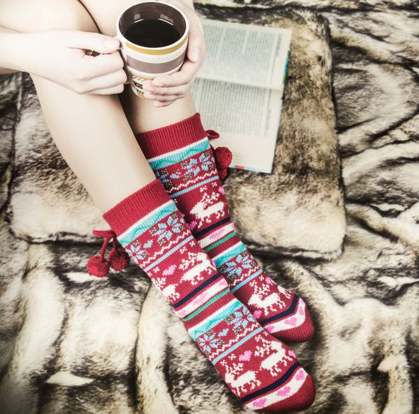 پاهای زن در جوراب های کریسمس با یک کتاب و یک فنجان قهوه خز مصنوعی روی زمین تصویر رنگ آمیزی شده