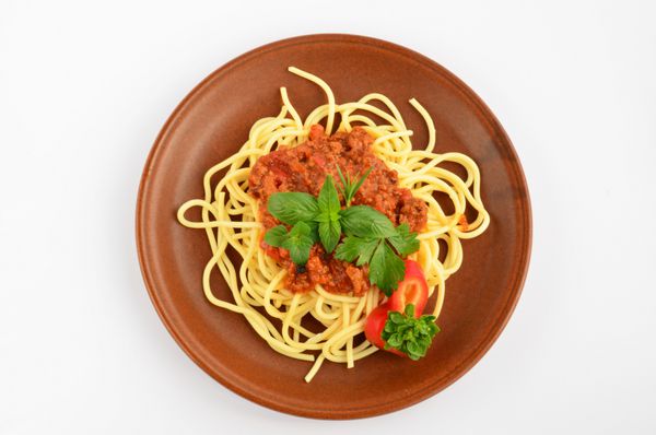 اسپاگتی بولونیز با ادویه‌های گیاهی تازه - پونه کوهی ریحان رزماری کرفس و فلفل قرمز در بشقاب سرامیکی قهوه‌ای روستایی روی زمینه سفید سرو می‌شود
