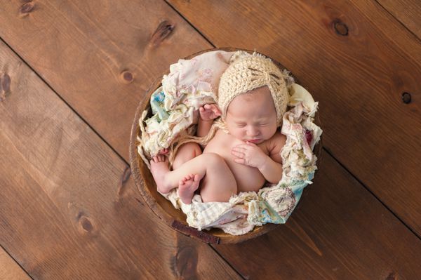 یک نوزاد دختر ده روزه که در یک کاسه چوبی کوچک می خوابد کلاهی به رنگ بژ بر سر دارد در استودیو روی پس‌زمینه چوبی