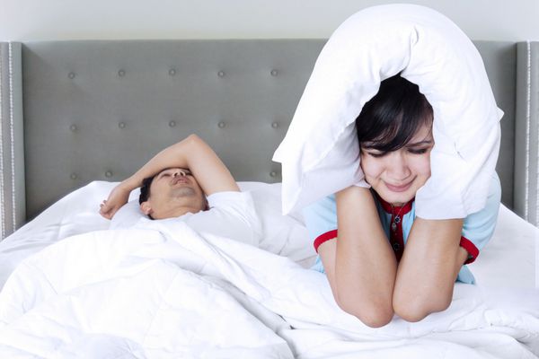 تصویر مرد جوانی که روی تخت خوابیده و خروپف می کند در حالی که همسرش نمی تواند بخوابد و از بالش برای پوشاندن گوش هایش استفاده می کند