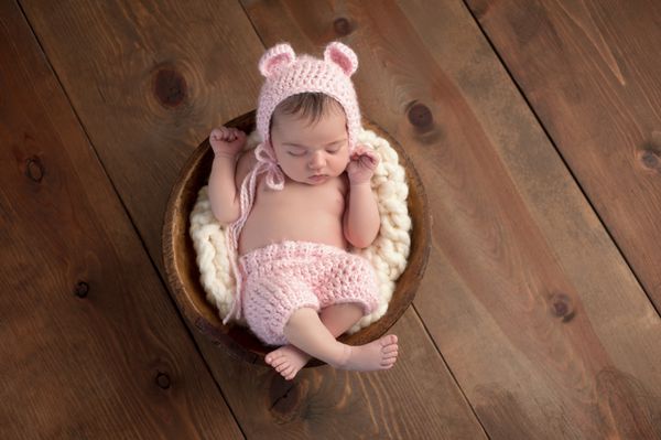 یک نوزاد دختر دو هفته ای که در یک کاسه چوبی کوچک می خوابد او یک کلاه خرس صورتی قلاب‌بافی و شلوارک همسان پوشیده است در استودیو روی پس‌زمینه چوبی