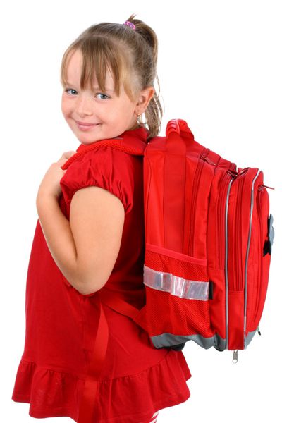 دختر کوچک با کیف مدرسه قرمز که به دوربین جدا شده روی سفید لبخند می زند