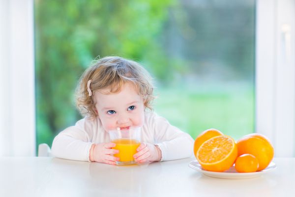 دختر نوپا ناز در حال نوشیدن آب پرتقال نشسته پشت میز سفید