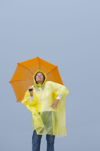 پرتره زن بالغ ایستاده زیر باران با پوشیدن بارانی زرد و چتر نارنجی جدا شده با آسمان خاکستری به عنوان پس زمینه و کپی sp
