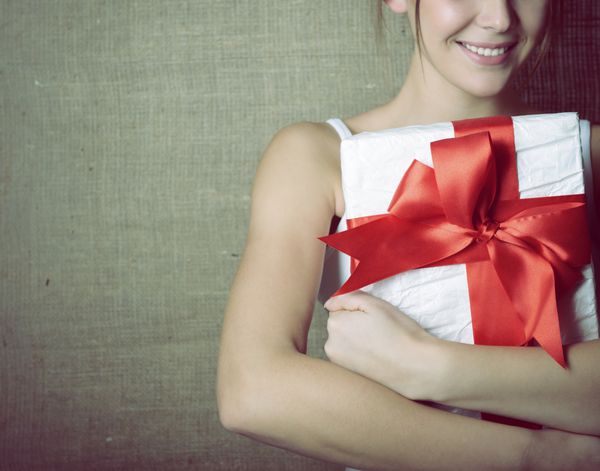 پرتره دختر شاد و جذاب با پیراهن سفید بدون آستین اسپرت که جعبه هدیه را در دست دارد با پاپیون قرمز روی پس زمینه بوم رنگ آمیزی شده و نویز اضافه شده است