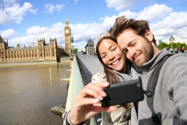 زوج توریستی لندن در نزدیکی بیگ بن در حال گرفتن پو هستند زن و مردی که در حال تفریح هستند با استفاده از دوربین گوشی های هوشمند لبخند می زنند و در نزدیکی دوست وست مینستر پل وست مینستر لندن انگلستان شاد می خندند