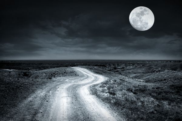 جاده روستایی خالی که در شب ماه کامل با آسمان ابری چشمگیر از میان دشت می گذرد