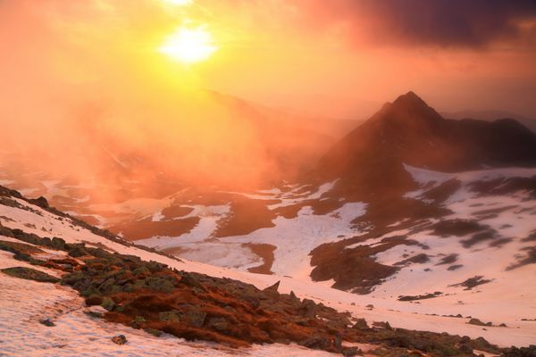 خورشید در هنگام غروب خورشید به ابرهای نارنجی بر فراز کوه برفی نفوذ می کند