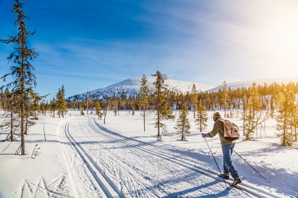 نمای پانوراما از مرد مرد در حال اسکی کردن در چشم انداز زیبای زمستانی شمال اروپا در اسکاندیناوی با آسمان آبی و نور طلایی عصر در غروب آفتاب اروپا