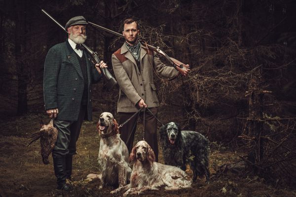 دو شکارچی با سگ و تفنگ در لباس تیراندازی سنتی در پس زمینه جنگلی تیره