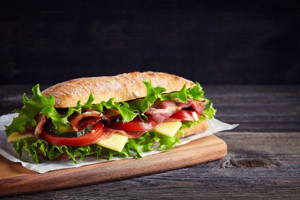 ساندویچ زیردریایی تازه با ژامبون پنیر بیکن گوجه فرنگی خیار کاهو و پیاز روی تخته برش چوبی
