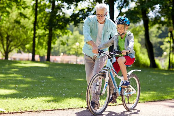 مفهوم خانواده نسل ایمنی و مردم - پدربزرگ خوشحال به پسر چگونه دوچرخه سواری را در پارک تابستانی آموزش می دهد
