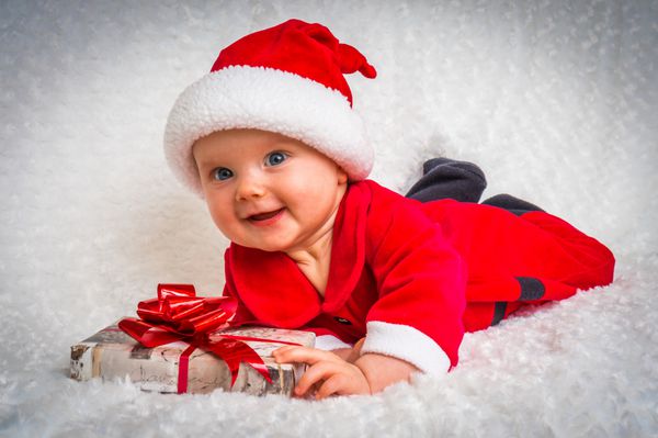 کودک خندان خوشحال که روی پتوی سفید با کلاه و کت و شلوار بابا نوئل دراز کشیده است - مفهوم کریسمس