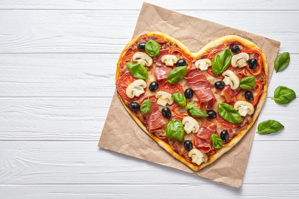 پیتزای شکل قلب خوشمزه مفهوم عشق روز رمانتیک رستوران شام غذای ایتالیایی زیتون پروشوتو شامپینیون گوجه فرنگی و موزارلا روی زمینه میز چوبی سفید سرو می شود