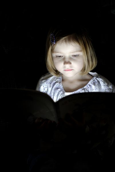 دختر کوچک در حال خواندن کتاب در تاریکی با نوری که بر روی f می تابد