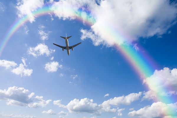 یک هواپیمای جت که بر فراز ابرهای سفید به سمت رنگین کمان در آسمان آبی پرواز می کند