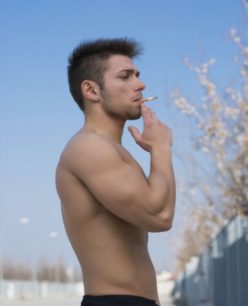 مرد جوان عضلانی بدون پیراهن در حال کشیدن سیگار در بیرون