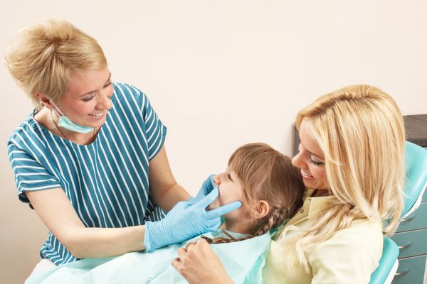 دختر بچه ای در آغوش مادر در حالی که دندانپزشک دندان های او را معاینه می کند