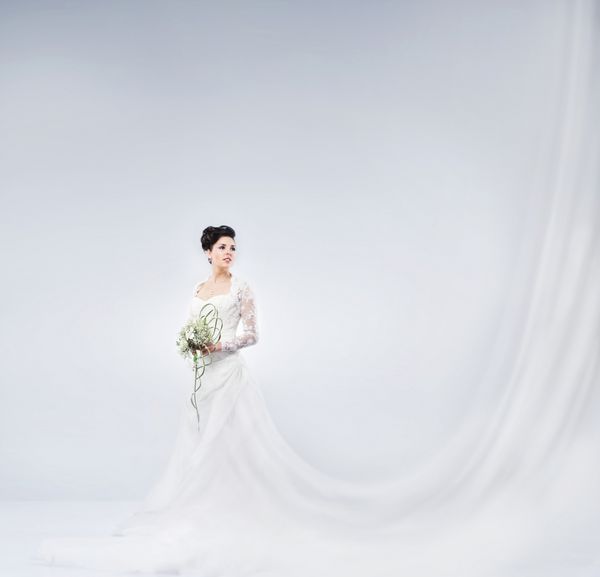 عروس جوان و زیبا در یک لباس فانتزی بلند ایستاده با یک دسته گل روی پس زمینه خاکستری