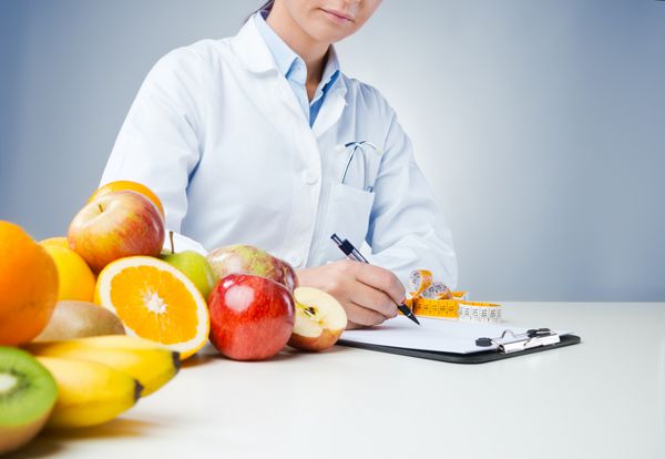 متخصص تغذیه حرفه ای که پشت میز کار می کند و سوابق پزشکی را با میوه های تازه در پیش زمینه می نویسد