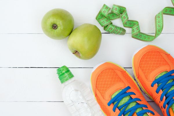 کفش ورزشی سیب و بطری آب در زمینه چوبی سفید تجهیزات ورزشی تمرکز انتخابی